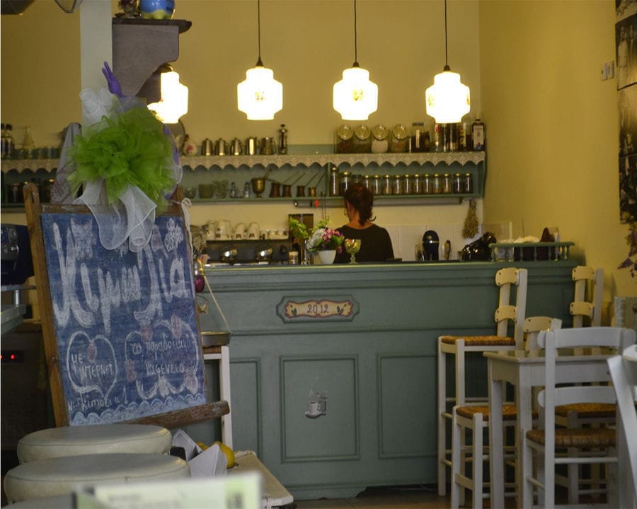 A charming Crete restaurant with a light blue wooden bar, white wooden chairs and tables, and a blackboard on the left. The restaurant's name is elegantly written in chalk, inviting guests to savor its culinary delights.